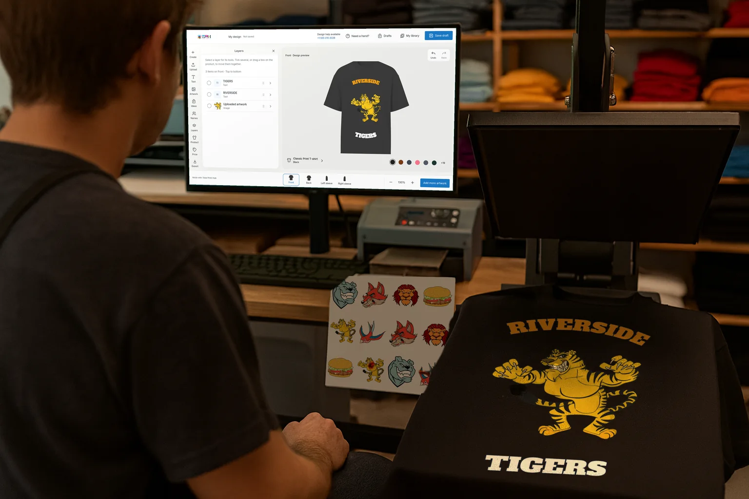 A print shop: a designer at a monitor showing the Total Print Hub Design Studio with the Riverside Tigers T-shirt, the same tiger design printed on a black T-shirt on the heat press, and a sheet of printed mascot transfers on the desk.